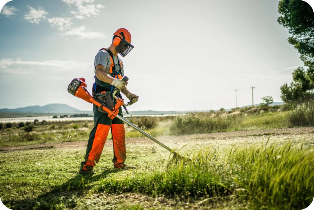 Worker using a string trimmer while wearing full PPE, including helmet, visor, gloves, and high-visibility gear.
