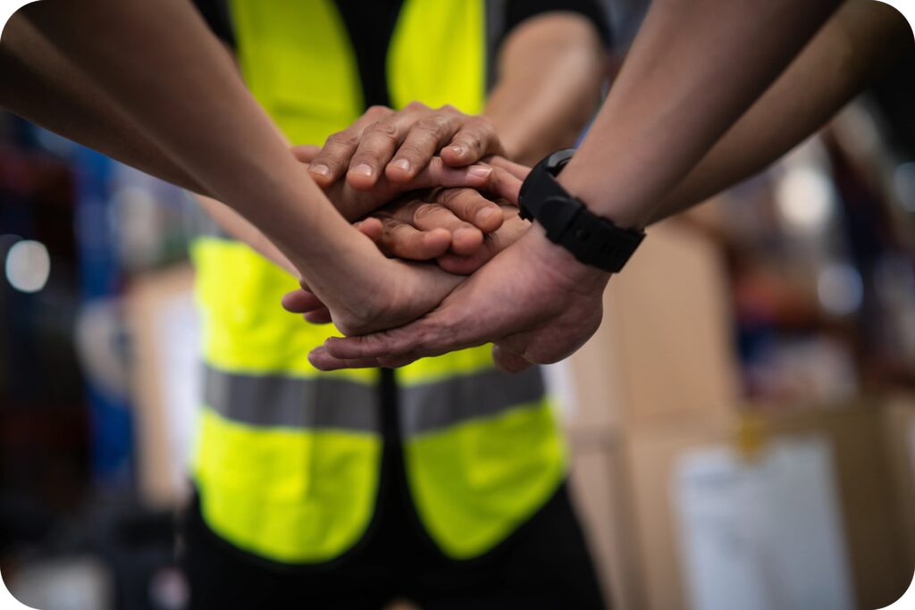 A group of workers in safety vests placing their hands together to show teamwork and collaboration in workplace safety.