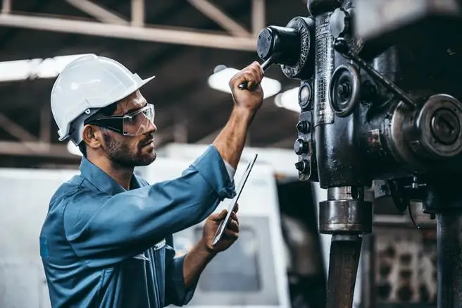 Technician in PPE inspect machinery while recording maintenance findings on tablet.
